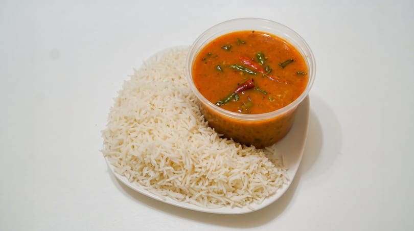 A clear plastic container of orange curry with green vegetables and red chilies, next to white rice.