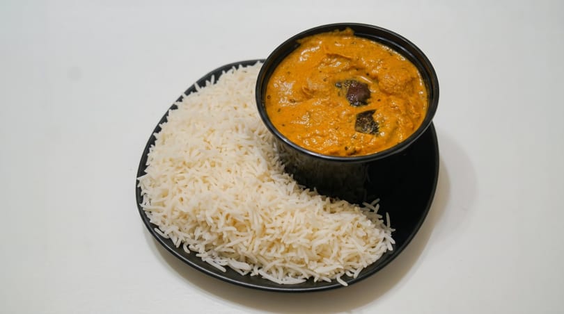 A black bowl of orange curry with dark spices next to a pile of white rice on a black plate.