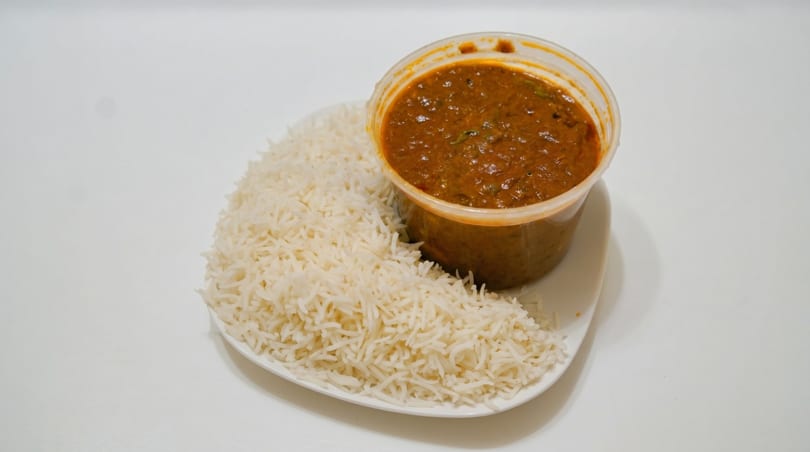 A clear plastic container of brown curry next to a pile of white rice on a white plate.