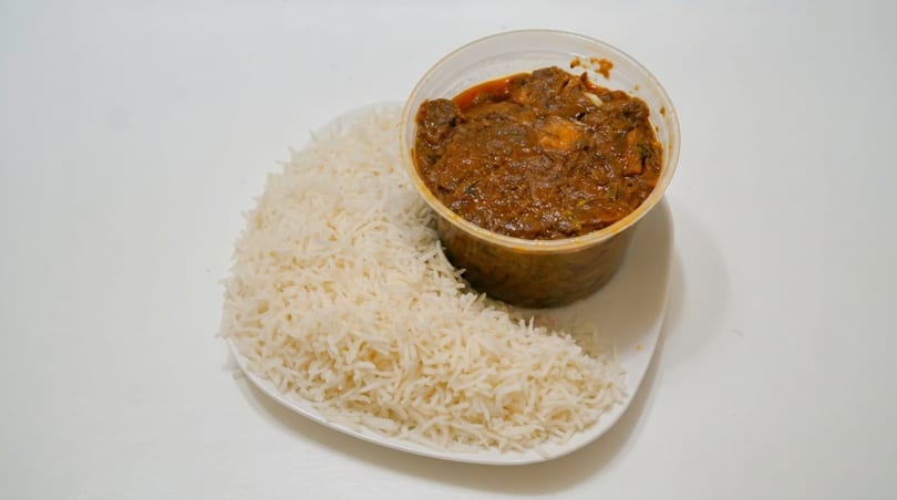 A clear plastic container of rich brown curry with white rice on a white plate.
