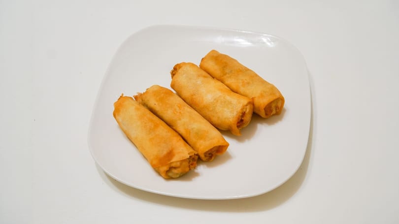 Four golden-brown spring rolls arranged on a white rectangular plate against a white background.