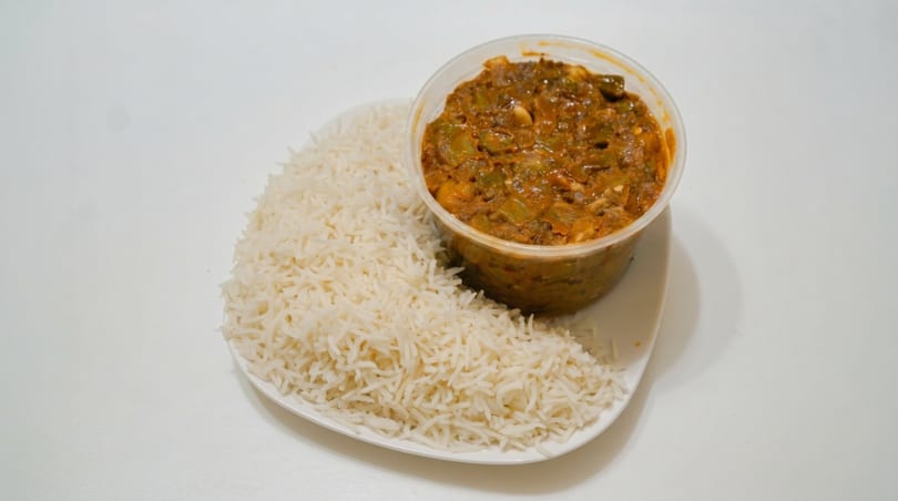 A takeout container of okra stew with a side of white rice on a white plate.