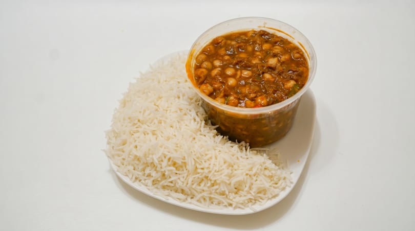 A white plate holds a mound of white rice next to a clear container of chickpea curry.